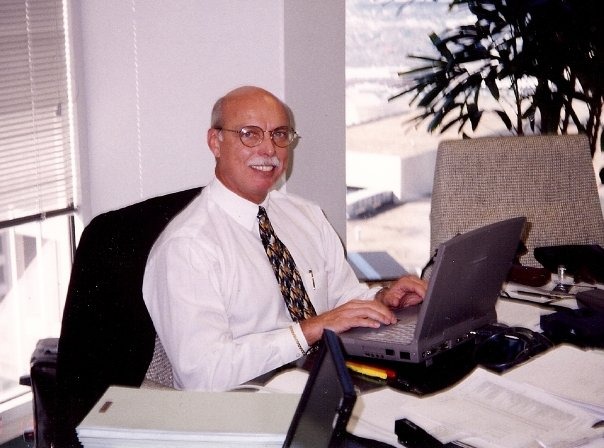 Bob McNeil in a shirt and tie, smiling over a laptop at a sunlit office desk