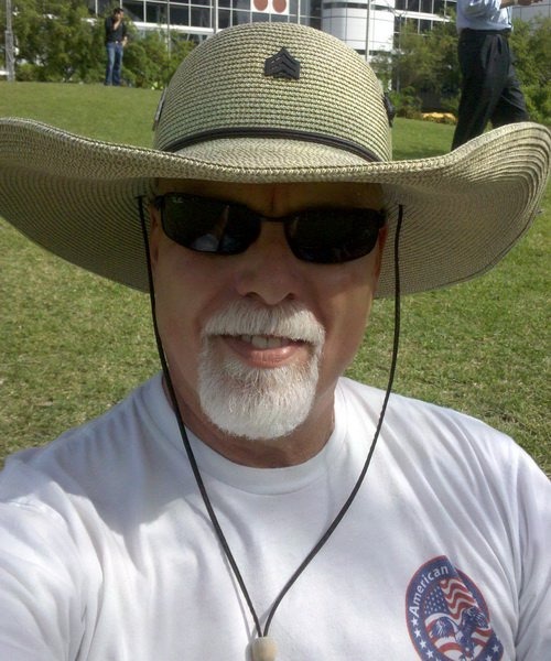 Bob McNeil outdoors in a wide-brimmed straw hat and sunglasses, white moustache and beard, on a sunlit lawn