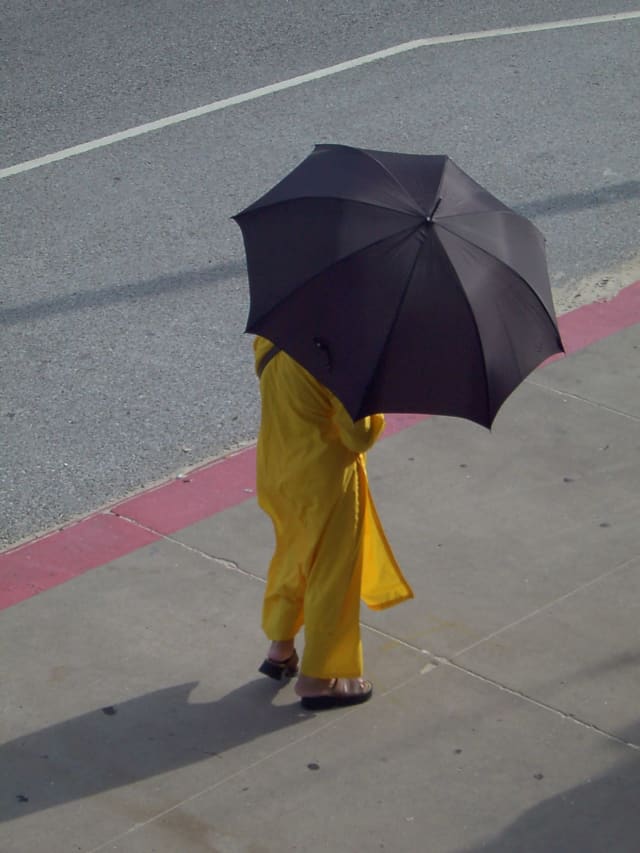A woman in a yellow dress crossing a sidewalk under a black umbrella, seen from above