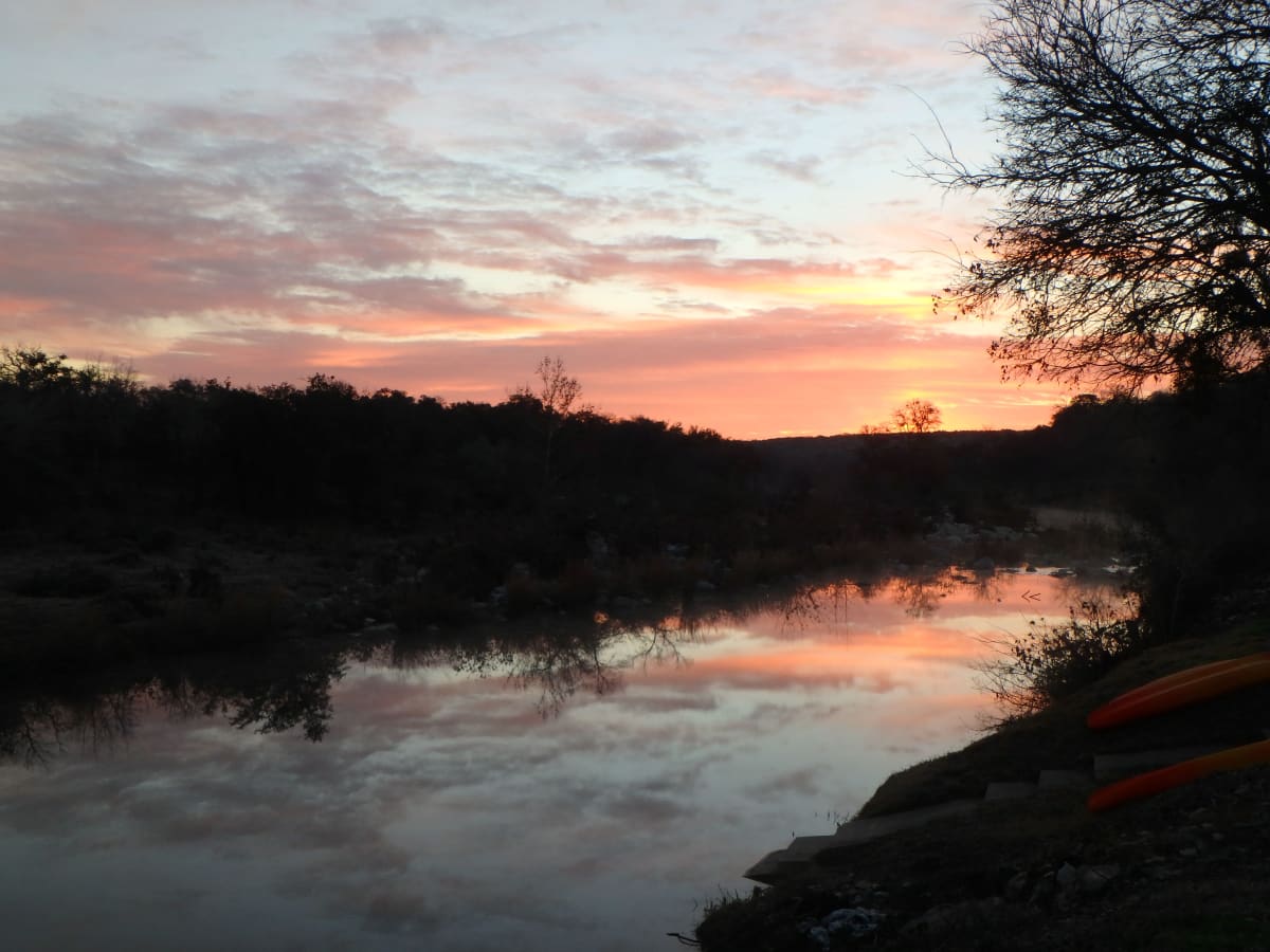 Pink and orange sunset reflected in a still Texas creek, kayaks pulled up on the far bank